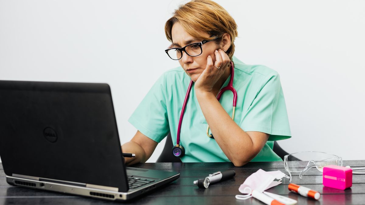 A tired clinician in scrubs reviewing records on a laptop